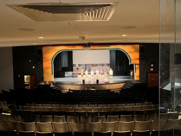 A group of six students stands on a theater stage, facing the audience in an empty auditorium with rows of black seats, a proscenium arch, and a projector screen, rehearsing under warm stage lighting.