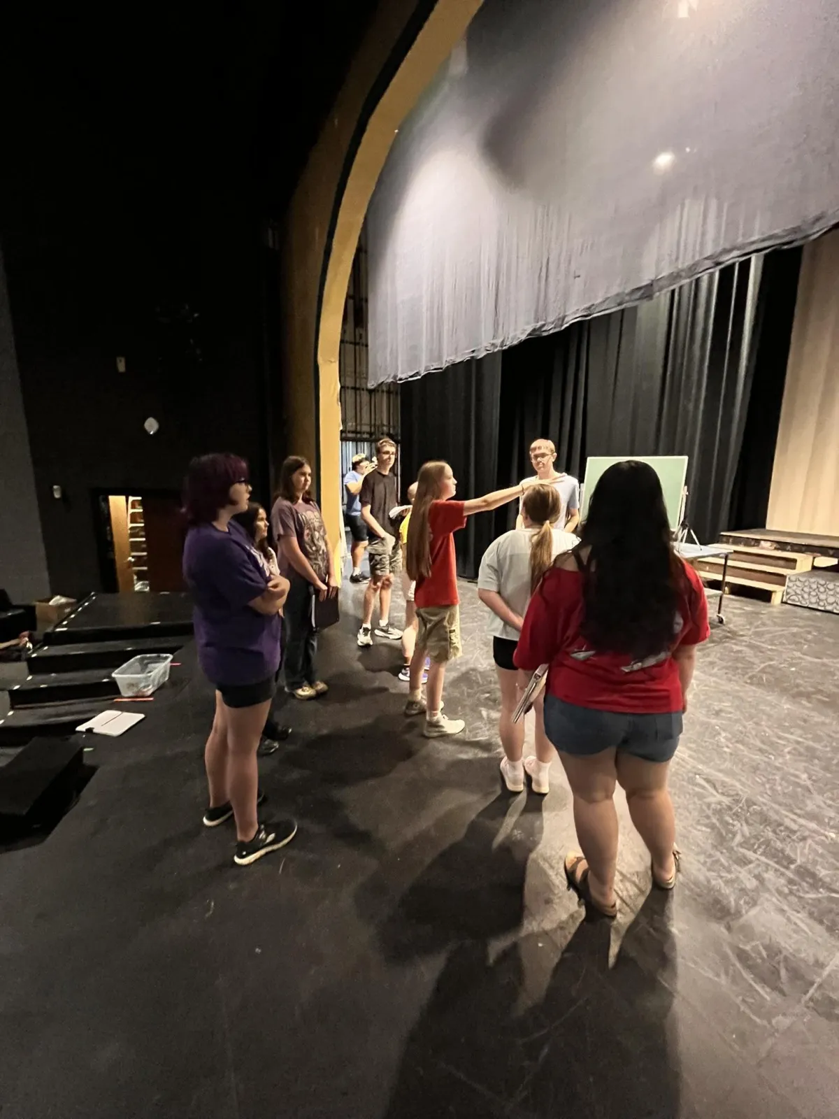 A group of students gathers on a theater stage with a black floor and draped curtains, collaborating and rehearsing under the warm glow of stage lights, with some gesturing and others listening intently.