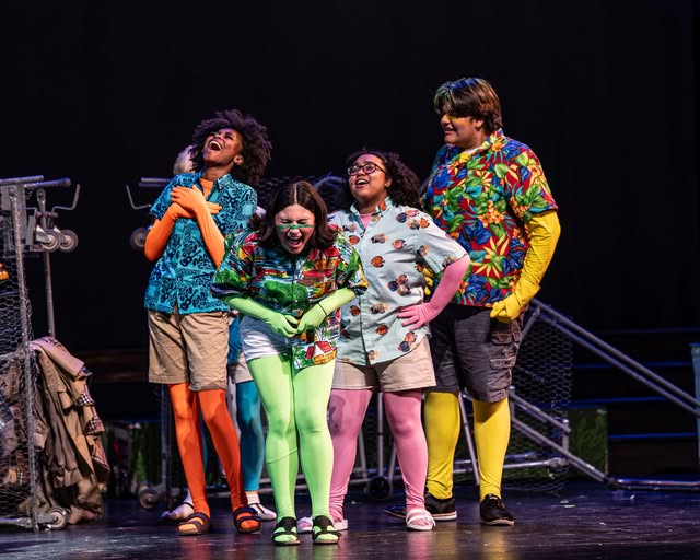 Four performers stand on a dimly lit stage, laughing and celebrating with bright, colorful costumes, including Hawaiian shirts, neon leggings, and mismatched accessories, exuding joy and camaraderie.