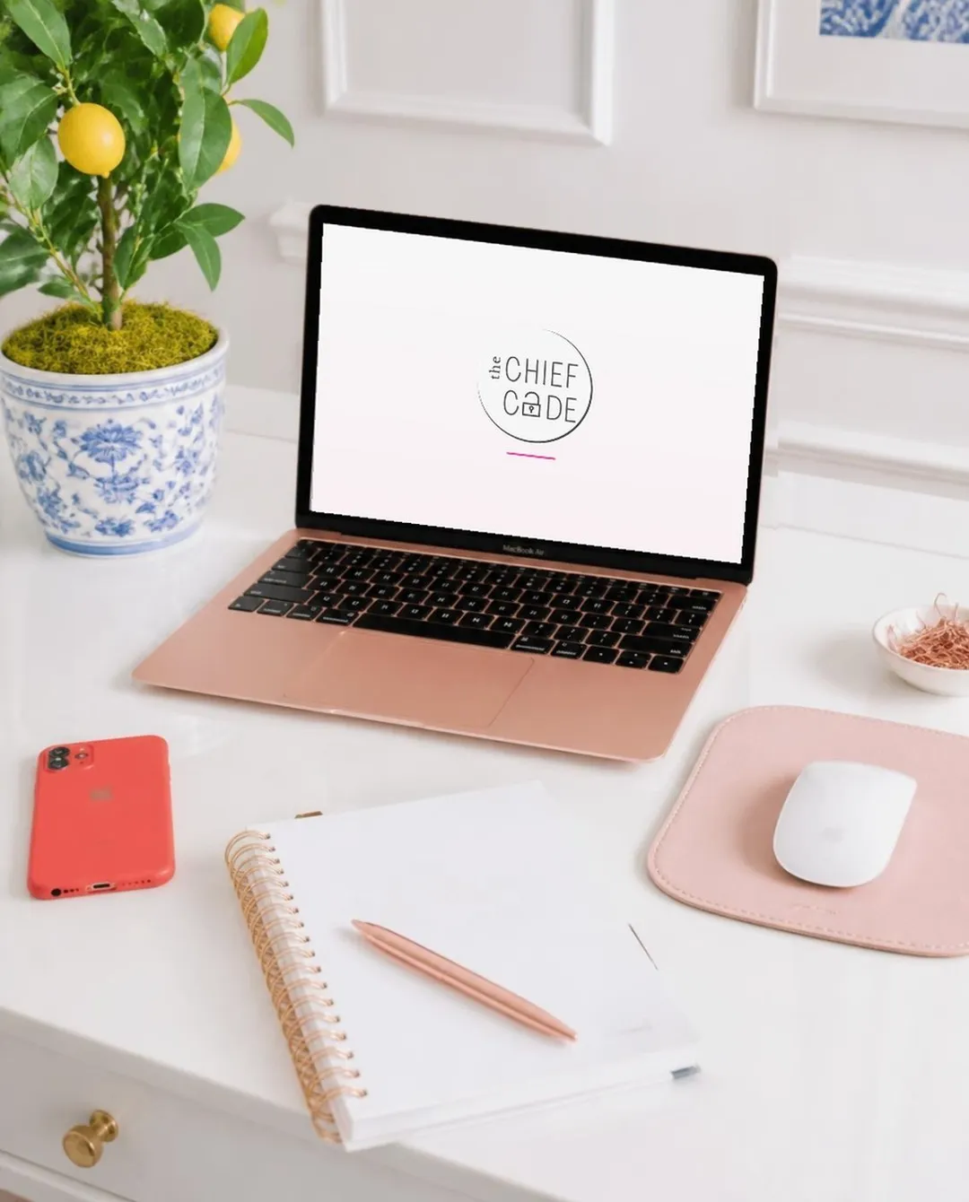 Rose gold MacBook showing The Chief Code logo on a white desk with a planner, pen, and lemon tree