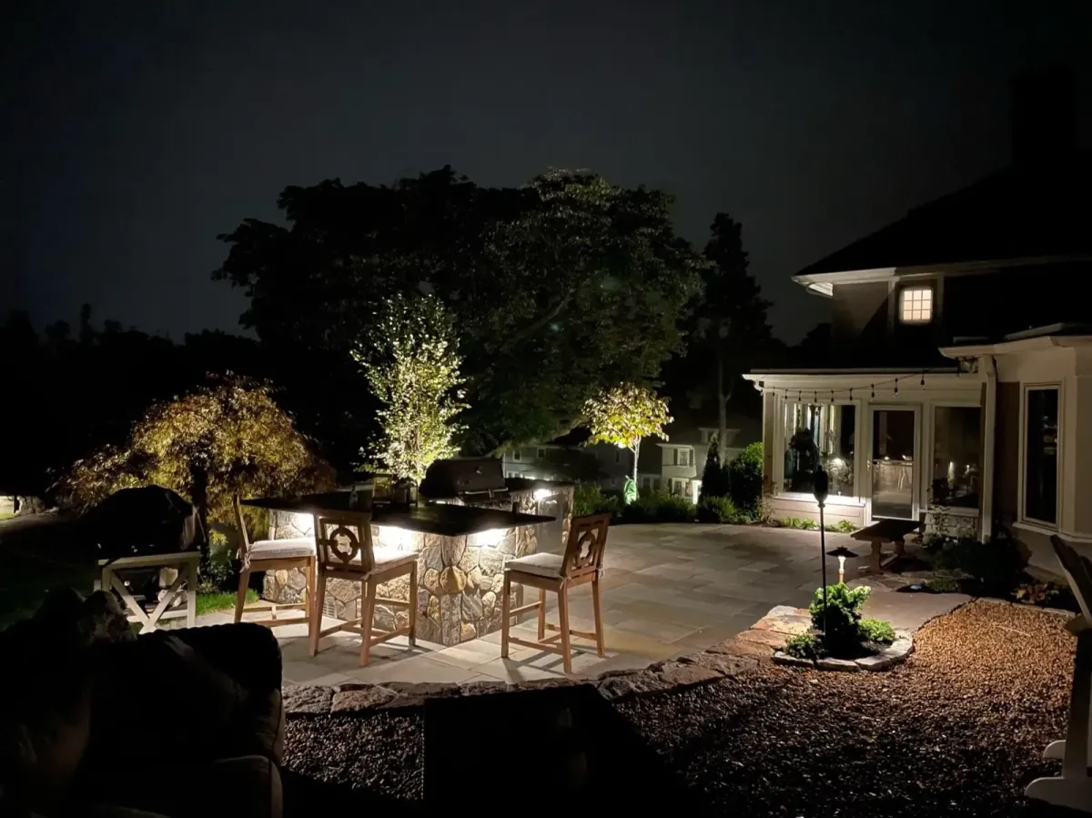 Illuminated backyard patio with custom stone outdoor kitchen, landscape lighting, mature trees, and professional landscaping in Marblehead, MA