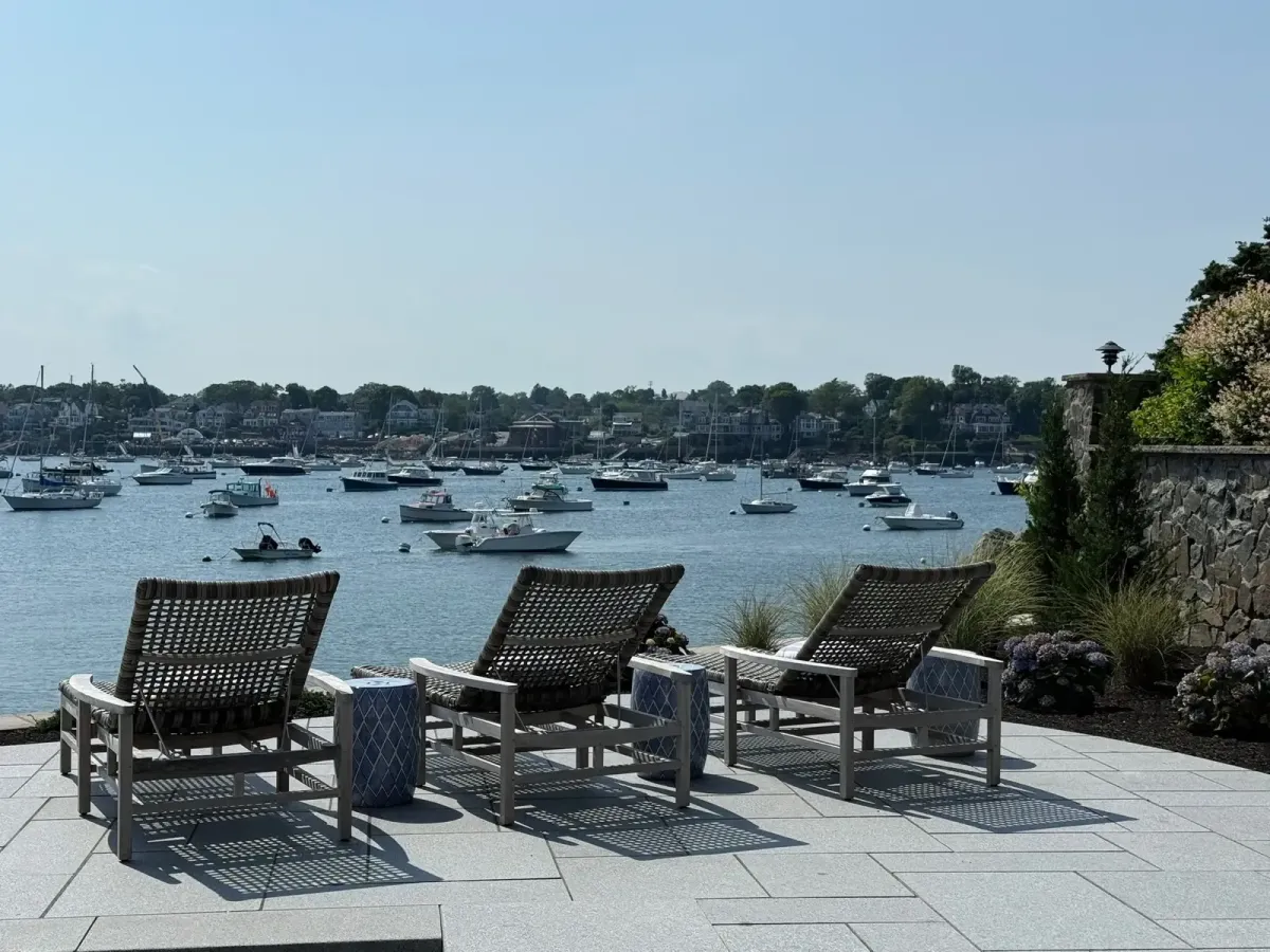 Waterfront stone patio with lounge seating, coastal landscaping, and panoramic views of Marblehead Harbor in Marblehead, MA