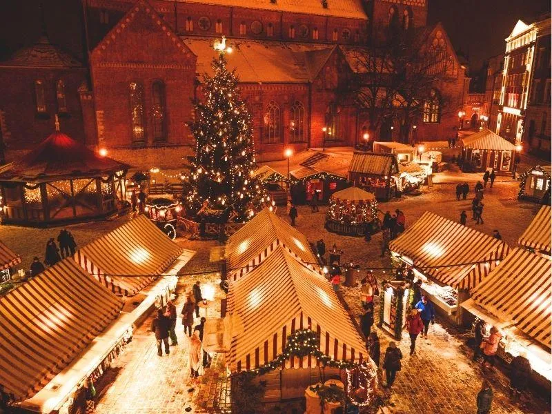 Nighttime scene of a festive European Christmas market with illuminated booths, a sparkling Christmas tree, and a church creating a magical atmosphere