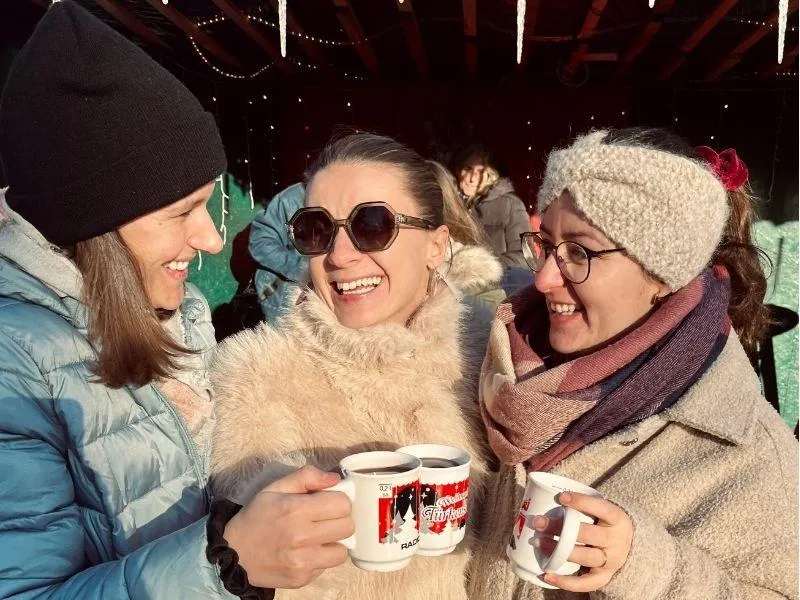 Three women sharing a cheerful toast with hot mulled wine at a Christmas market, surrounded by holiday lights