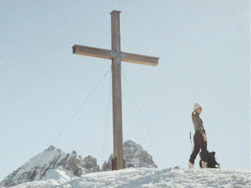 Summit cross on top of a mountain with a skier standing next to it, showcasing strength and achievement against a clear alpine backdrop