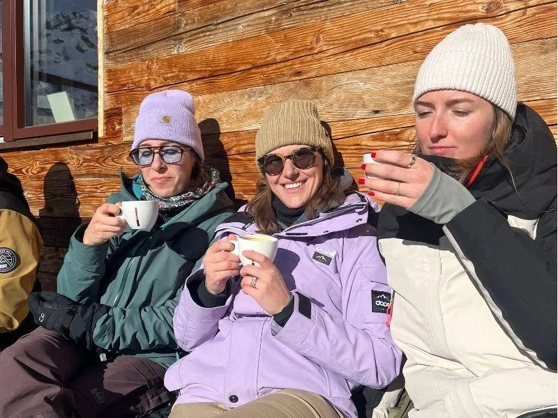 Three women in ski gear sitting in front of a mountain ski hut, wearing sunglasses and enjoying coffee in the sunny Alps