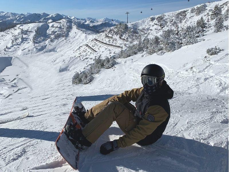 Snowboarder wearing goggles, helmet, and face mask sitting on a mountain peak with snowy slopes and stunning alpine scenery in the background