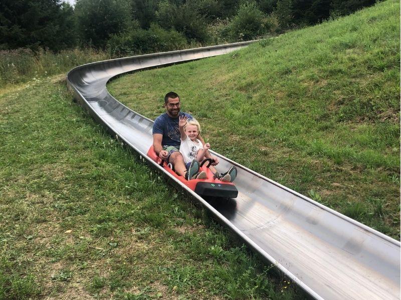 Adult and young girl riding a summer sledding bob track in Germany, with the child smiling and waving at the camera during a fun family adventure