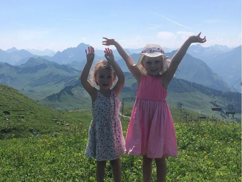Two young children raising their arms joyfully in front of a scenic mountain panorama in Switzerland during a family travel adventure
