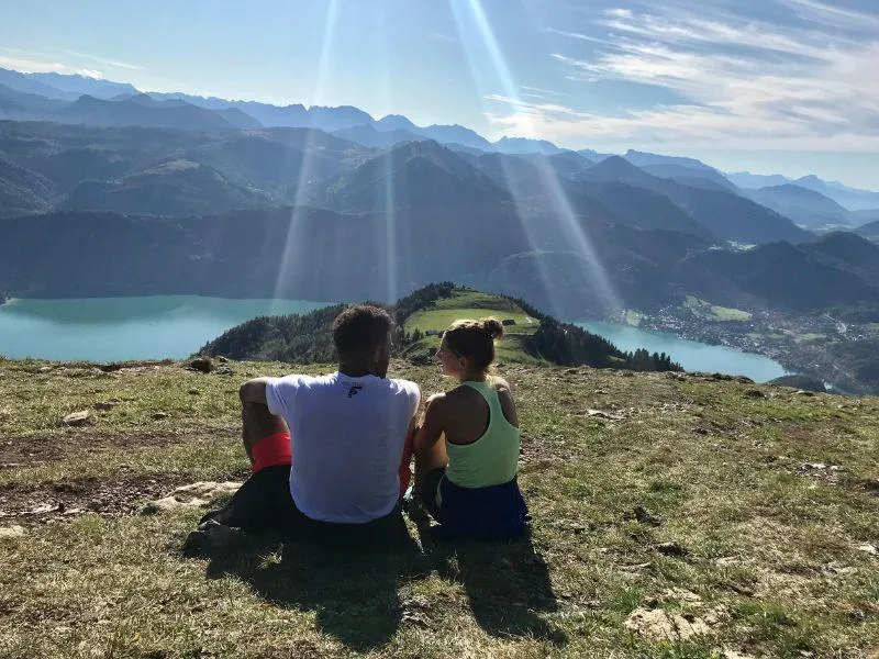 Couple sitting in a grassy alpine meadow overlooking panoramic mountain views and a lake in the Alps