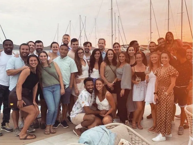 Large group of friends posing for a photo with sailboat masts and a marina in the background during a European trip