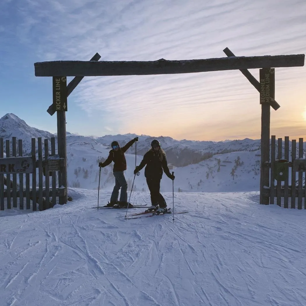 Two skiers gliding down a snowy mountain with scenic peaks and a glowing winter sky”