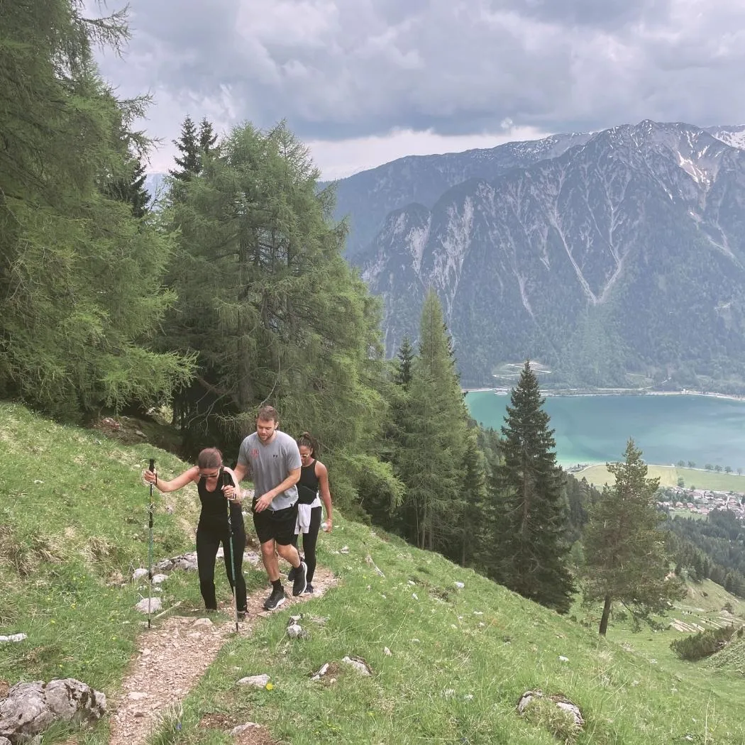 Three hikers trekking a mountain trail with walking sticks, overlooking trees, a train, lake, and distant Alps