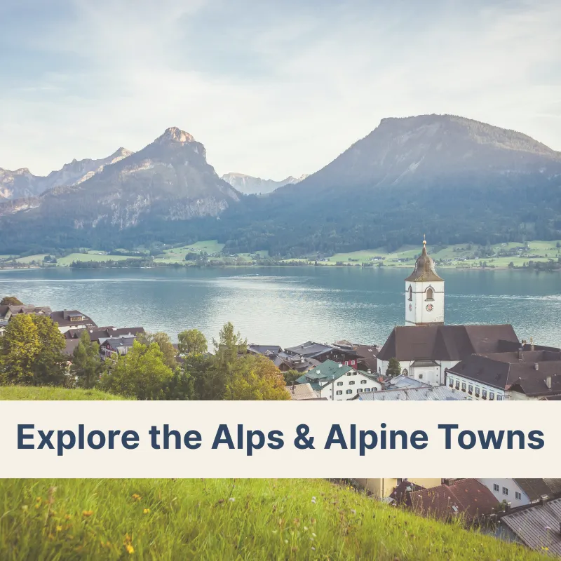Alpine town with a church and colorful buildings in a valley, a lake in front, and majestic mountains surrounding it, representing Alps & Alpine Towns tours.