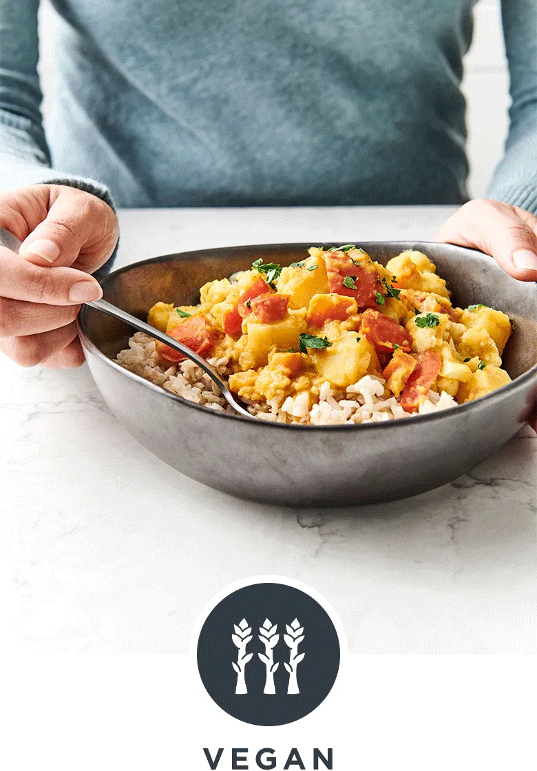 Woman eating a vibrant hearty vegan curry in a black ceramic bowl on a white marble countertop
