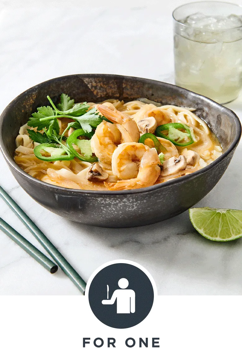 Black ceramic soup bowl filled with succulent shrimp pho with lime wedge, chopsticks & a glass of water on a white marble surface