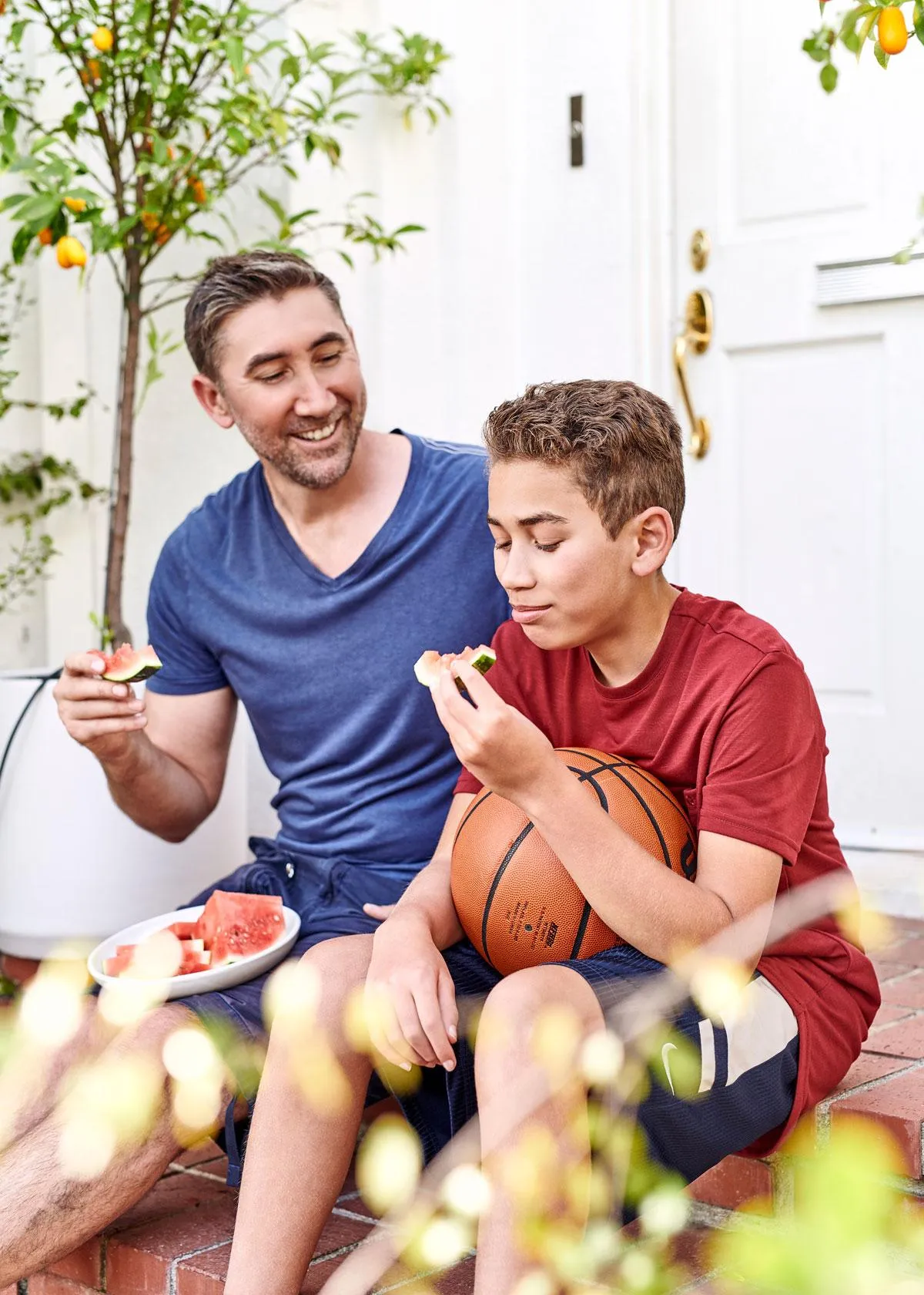 Father and son enjoy fresh watermelon on a porch, smiling and bonding. The boy holds a basketball. Healthy eating, family time.