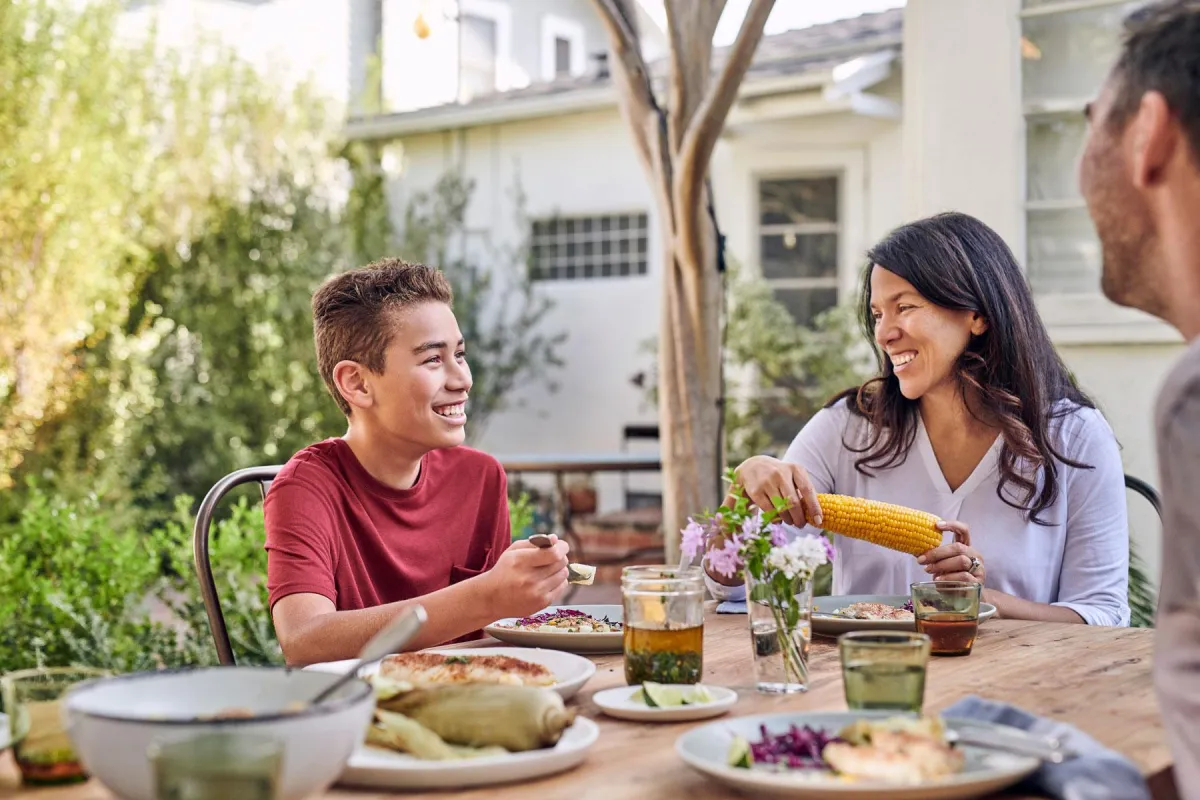 Smiling family enjoys an outdoor meal with fresh food, including corn and tamales, on a wooden table in a sunlit backyard.