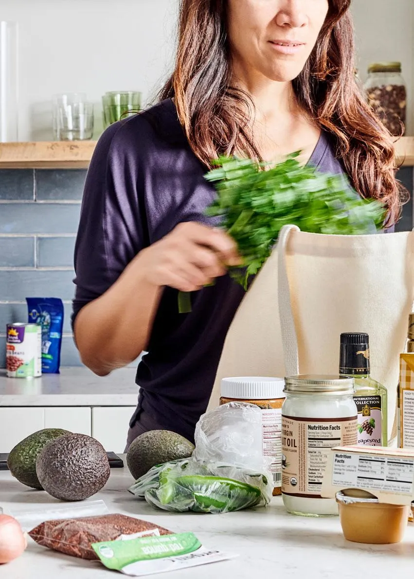 Woman unpacks fresh groceries, including avocados, greens, and pantry staples, onto a kitchen counter for healthy meal prep.