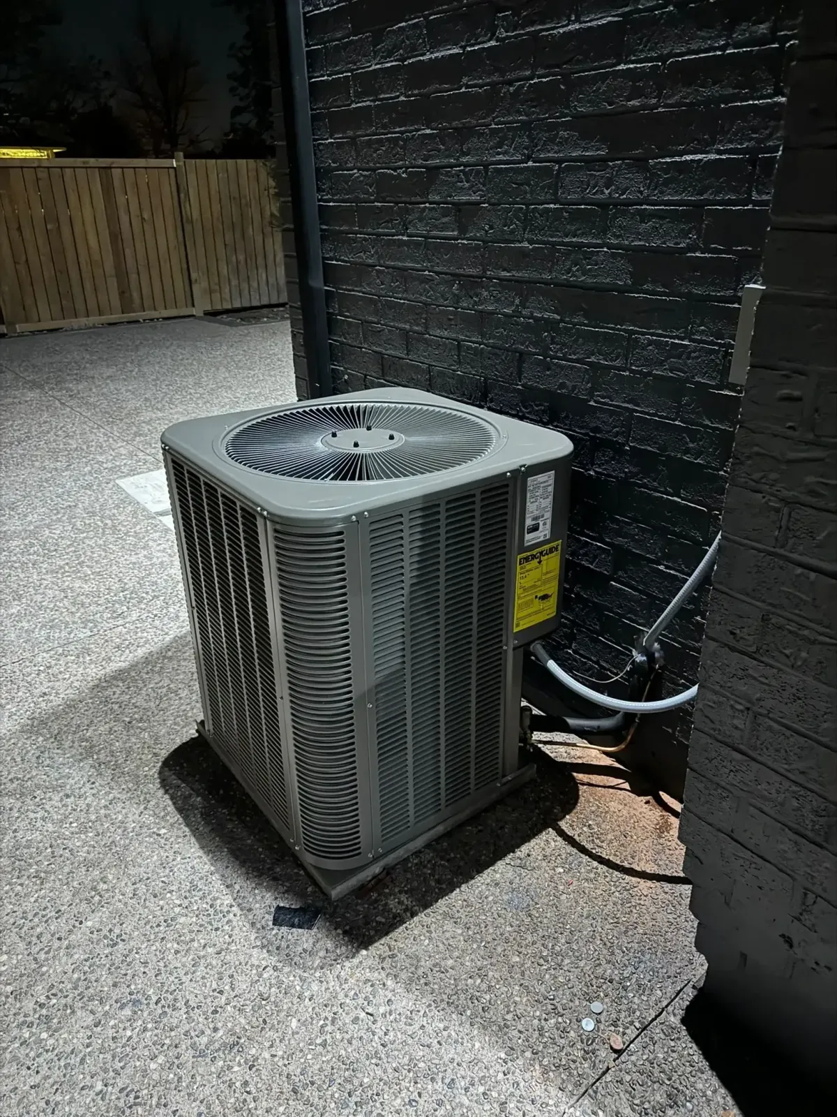 Outdoor air conditioning unit against a dark brick wall with warning label, cables, and wooden fence in dim light.