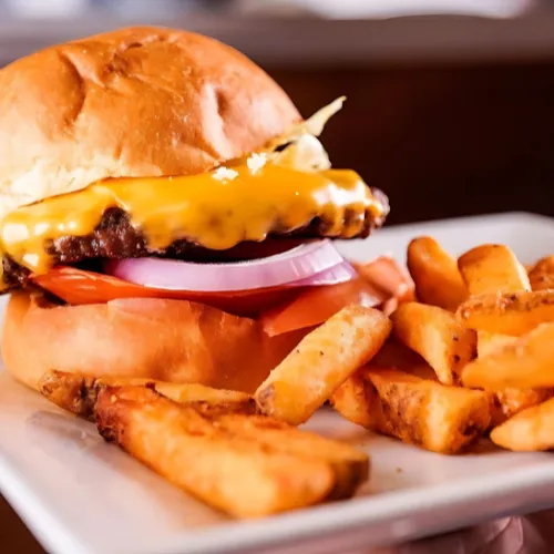 Juicy cheeseburger with melted cheddar, red onions, tomatoes, and lettuce on a toasted bun, served with crispy fries on a white plate.