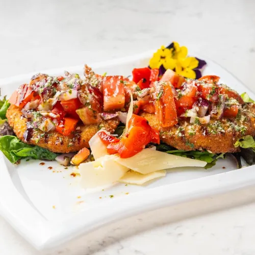  plate of bruschetta topped with diced tomatoes, shredded cheese, and balsamic glaze, served on a white square plate with fresh greens and a yellow edible flower.