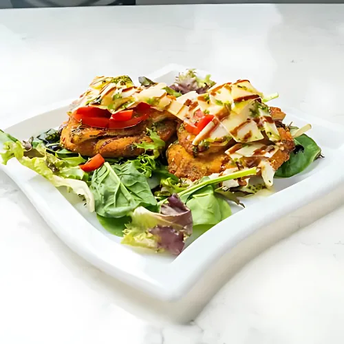 A fresh salad with crispy breaded protein, diced tomatoes, and a drizzle of dressing, served on a bed of mixed greens on a white square plate.