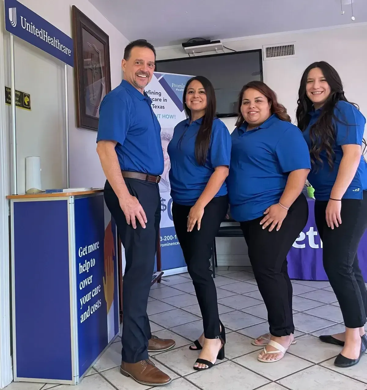 A group of four professionals, dressed in blue polo shirts and black pants, stand together next to a UnitedHealthcare display.