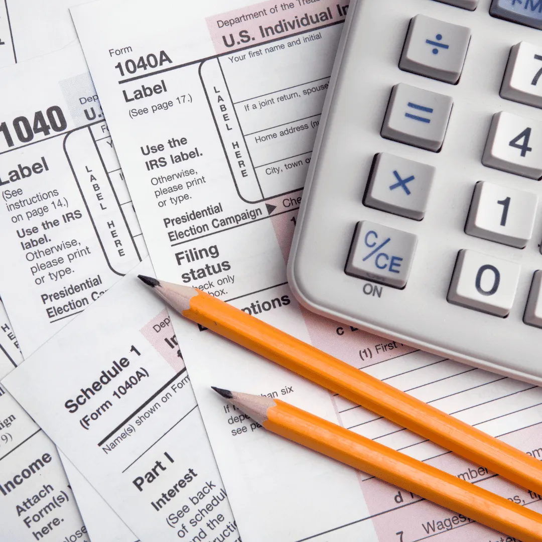 A close-up of IRS tax forms (1040 and Schedule 1) with a calculator and two pencils.