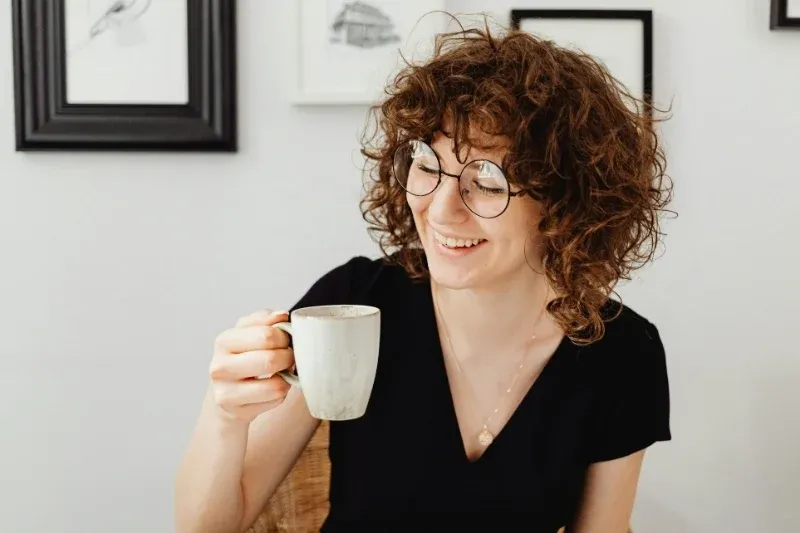 Woman with curly hair and glasses holding a white mug, smiling, sitting indoors.