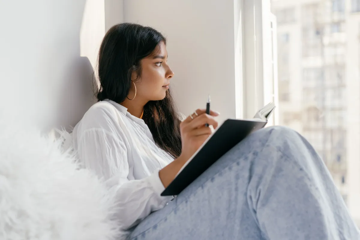 Woman sitting comfortably near a window, writing or reading on a tablet or notebook.