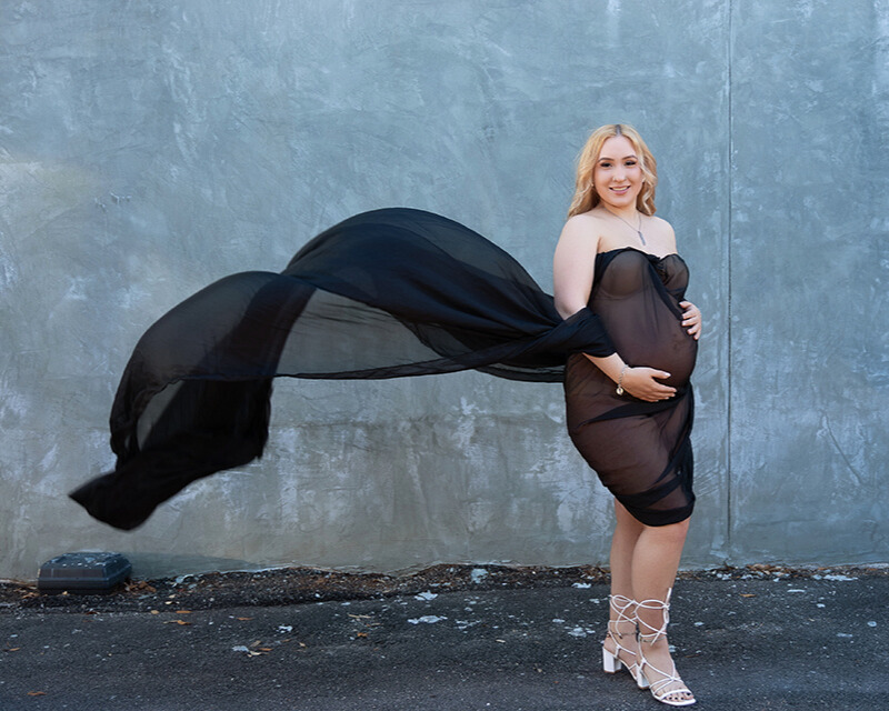 A pregnant woman in a flowing white dress stands in a sunlit field, gently cradling her belly. The golden hour light creates a soft, ethereal glow, and wildflowers add a touch of color to the serene landscape.