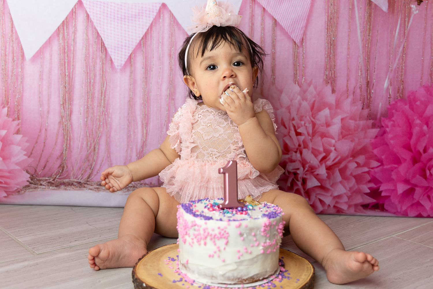 A joyful toddler sits on a pastel-colored floor, hands covered in cake and frosting, with a partially smashed cake in front. Balloons and streamers in the background add a festive, playful atmosphere.