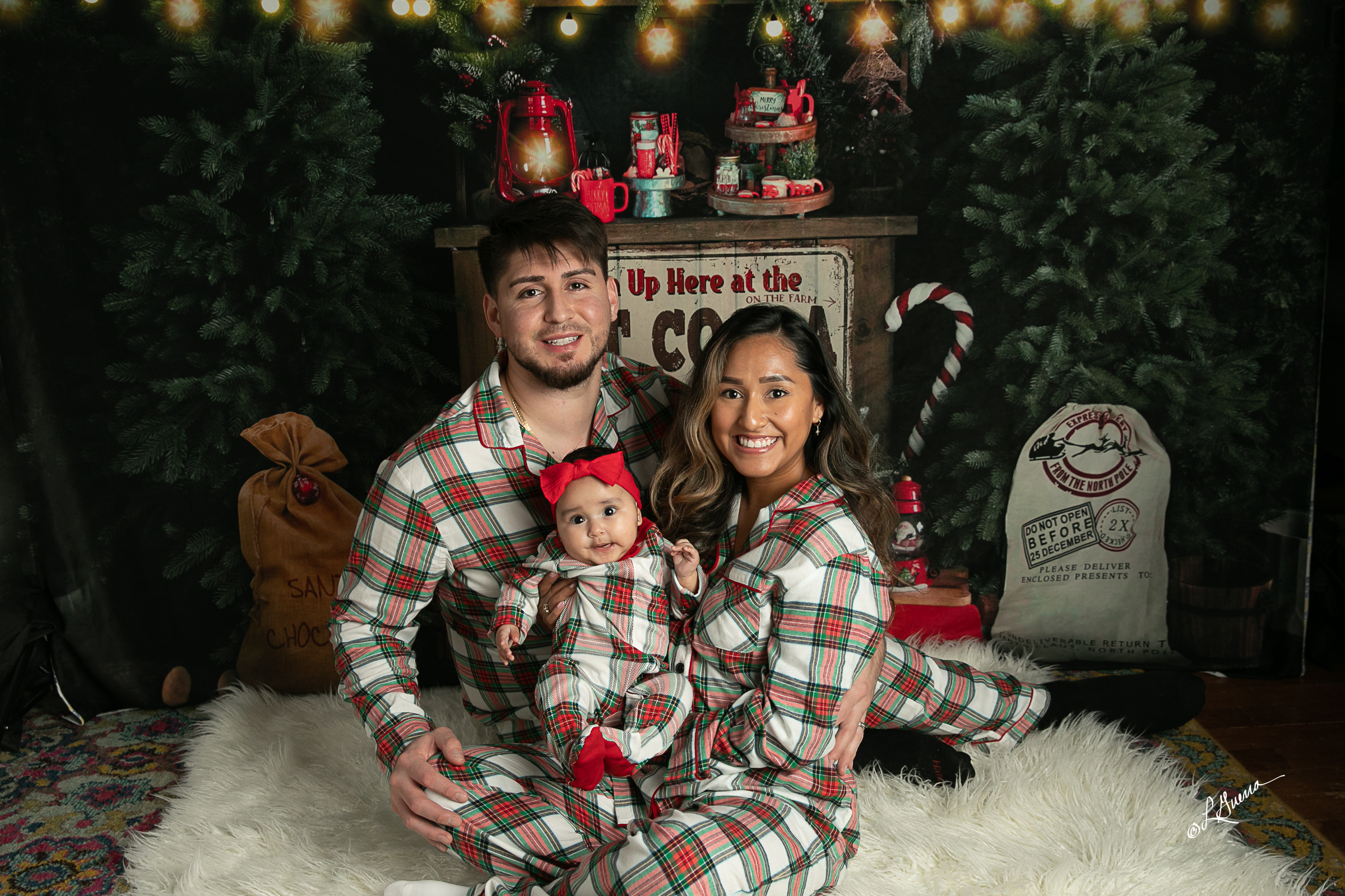 A couple and their young child, all in coordinated red and green outfits, sitting on a simple bench with a modern Christmas tree and wrapped gifts in the background. The set is bright, uncluttered, and festive.