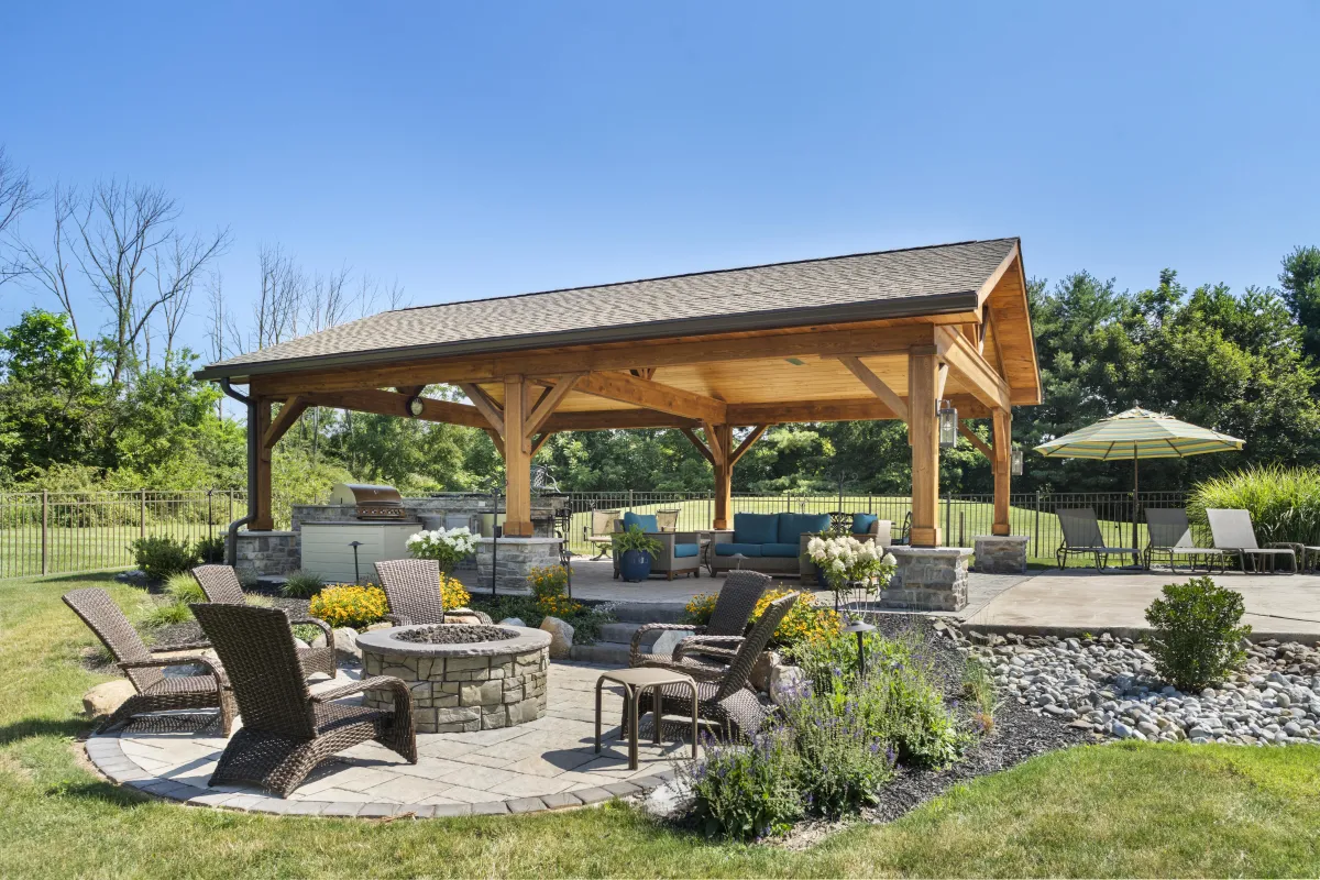 Beautiful outdoor living space featuring a custom-built wooden pavilion, a cozy fire pit with surrounding wicker chairs, an outdoor kitchen, and lush landscaping—perfect for relaxation and entertaining.