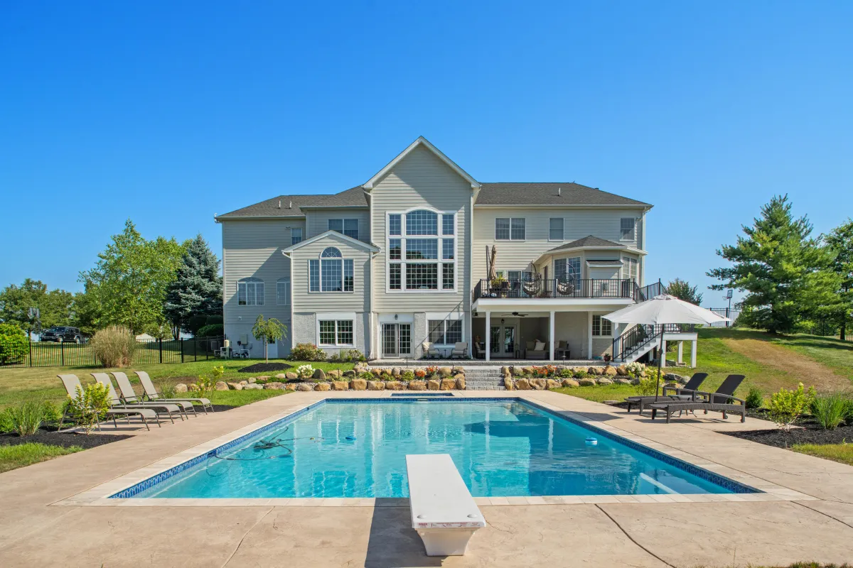 Luxury backyard with a custom in-ground pool, surrounded by a spacious patio, lounge chairs, and lush landscaping. A beautiful three-story home with a covered porch and elevated deck overlooks the pool, creating a perfect outdoor living space for relaxation and entertaining.