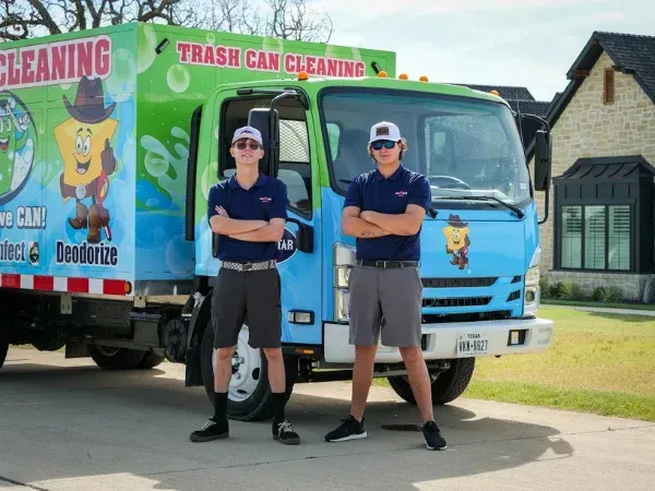 Two young men in uniform with crossed arms standing proudly in front of a blue and green trash can cleaning truck.