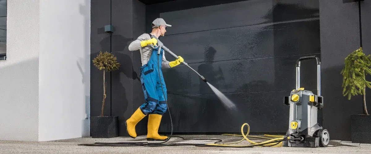 A man in blue uniform pressure washing two trash bins with a colorful service truck in background.