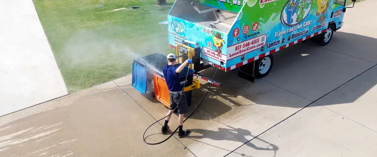 A man in a blue shirt and white hat pressure washing a blue trash can on a driveway next to a service truck.