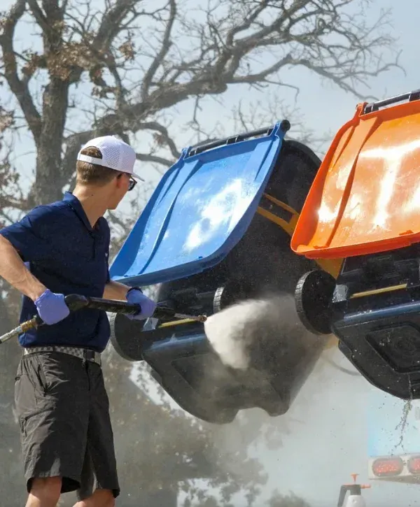 A man in shorts and a white hat power-washing hanging blue and orange recycling bins under a tree.