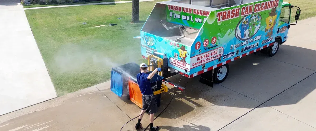 Two young men in uniform stand proudly next to a vividly branded trash can cleaning truck in a suburban setting.