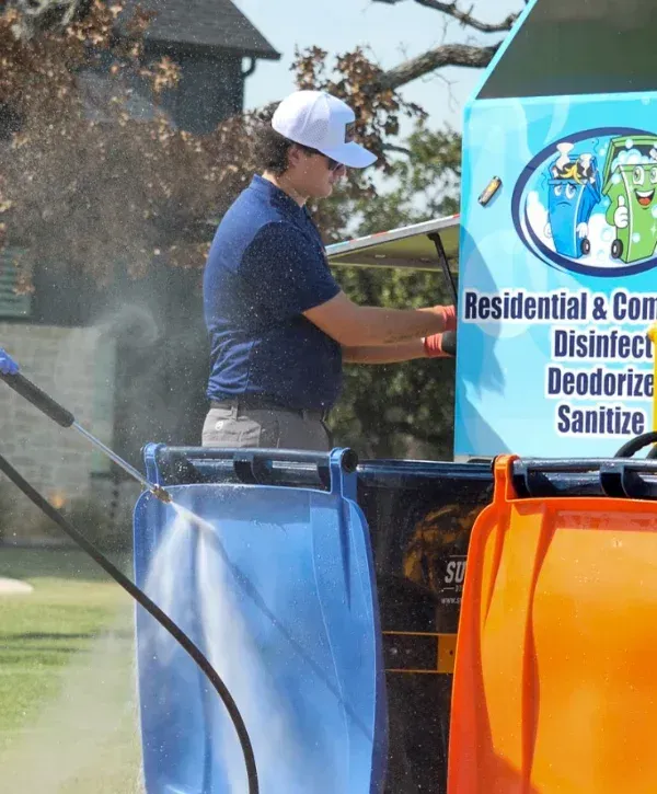 A man in a white cap and blue polo shirt pressure washing a blue trash bin outdoors on a sunny day.