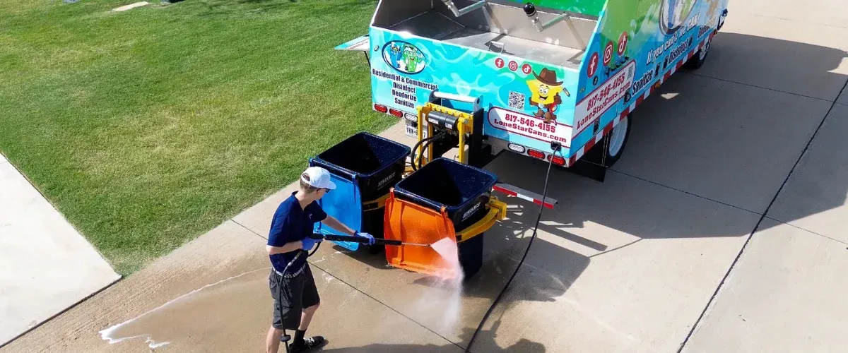 A man in blue uniform power washing an orange trash bin, next to a blue garbage cleaning truck.