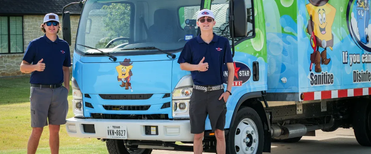 A man in blue uniform power washing an orange trash bin, next to a blue garbage cleaning truck.