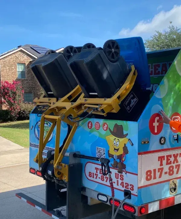 Rear view of a blue and green trash truck with two black bins elevated and tilted for emptying, revealing colorful decals and a pressure washer nozzle.
