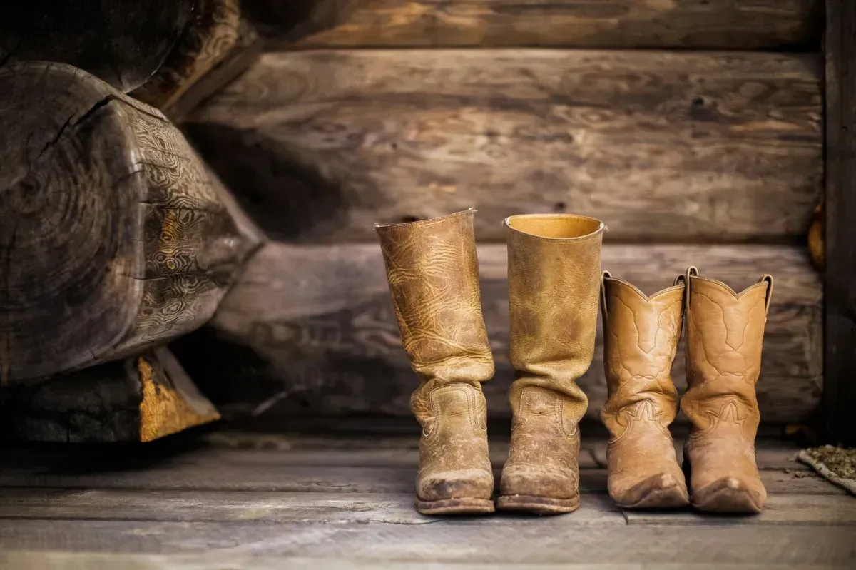 Two pairs of worn leather cowboy boots resting on a rustic log cabin porch in Powell Wyoming — authentic Western lifestyle homes for sale with Wild West Real Estate Group