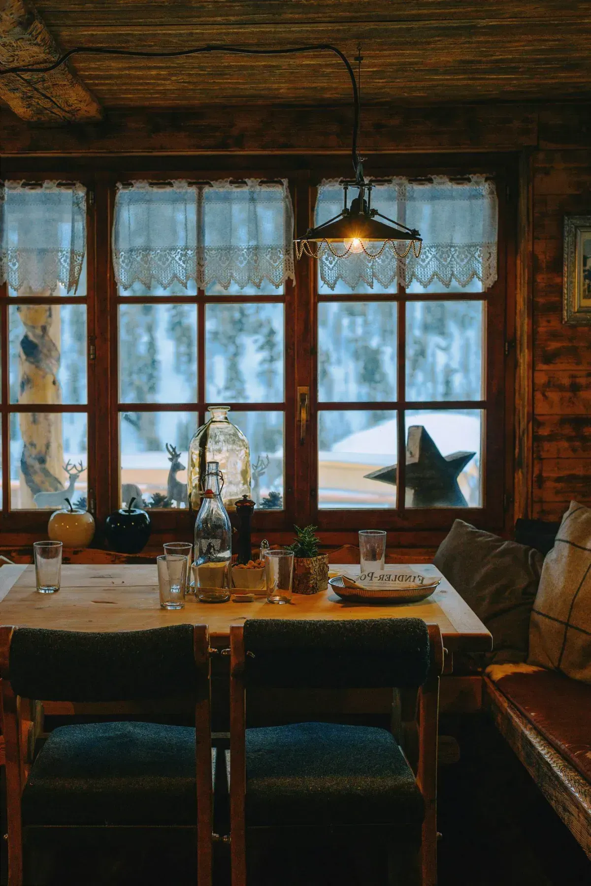 Warm rustic log cabin interior with wooden dining table, pendant light, lace curtains, and snowy winter forest view — cozy home for sale in Lovell Wyoming Big Horn Basin