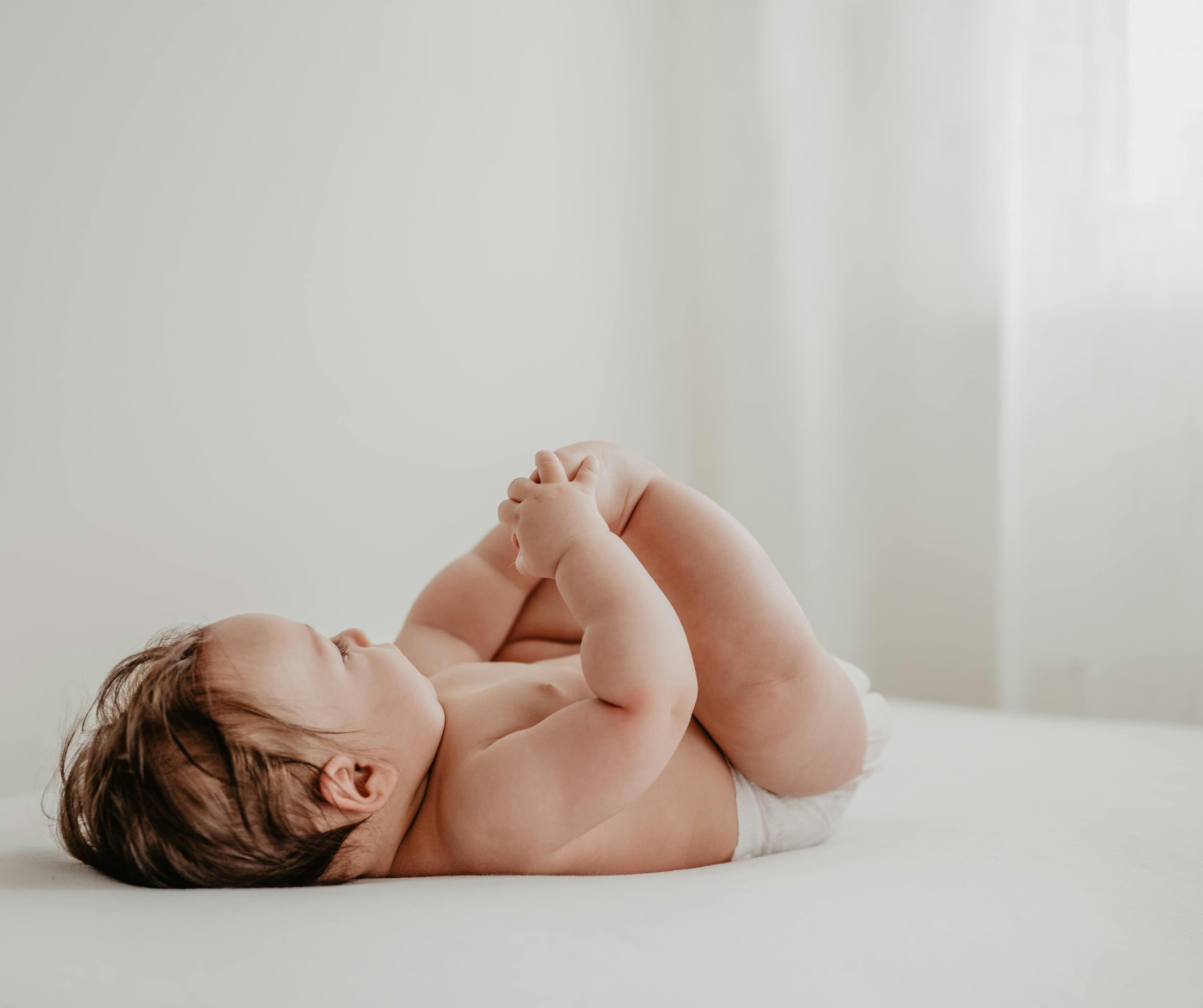 Baby lying on a white bed, holding their feet in a softly lit nursery.