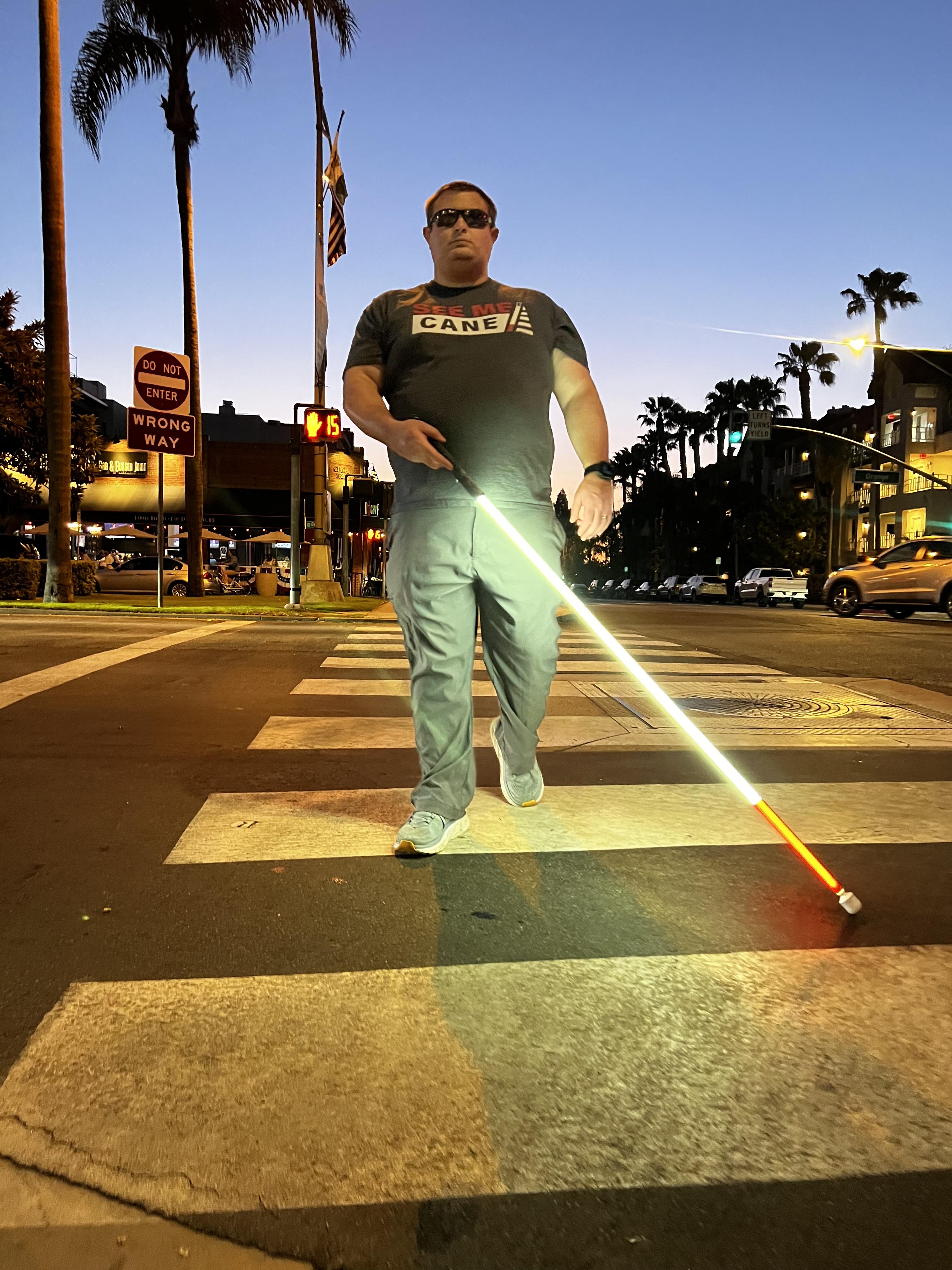 A blind pedestrian crossing a street at dusk using a glowing lighted white cane