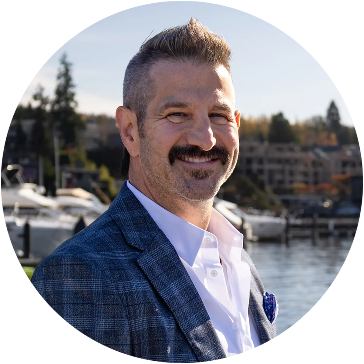 Jason Stricevich smiling outdoors on a sunny day in Kirkland, standing near the waterfront with boats and modern homes in the background. Dressed in a blue plaid blazer and white shirt, he presents a friendly and professional appearance.